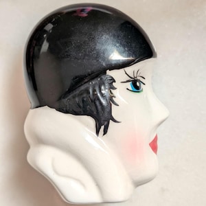May include: A ceramic figurine of a woman's head in profile. The head is painted with a black cap, black hair, blue eyes, red lips, and pink cheeks. The figurine has a white collar and is likely a decorative item.