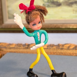 May include: A vintage bendable doll with brown hair and a red bow. The doll has a green torso, yellow legs, white hands, and black shoes. The doll is posed with one arm raised and the other extended.