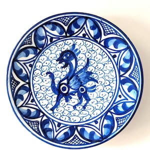 May include: A round ceramic plate featuring a hand-painted blue and white design. The center displays a stylized bird figure surrounded by small circular patterns, while the border is decorated with repeating leaf-like shapes and cross-hatching.