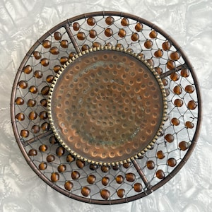 Puede incluir: Una bandeja decorativa redonda de metal con un centro martillado de color cobre. La bandeja está rodeada por una malla de alambre y adornada con cuentas de color ámbar. El borde exterior es de un color bronce más oscuro. La bandeja es decorativa.