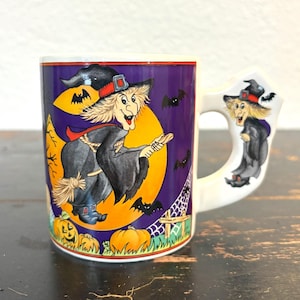 以下が含まれることがあります： 白いセラミックハロウィーンマグ。漫画の魔女が三日月を背景にほうきに乗っています。マグカップは、コウモリ、カボチャ、ハンドルに魔女がいる紫色の背景です。