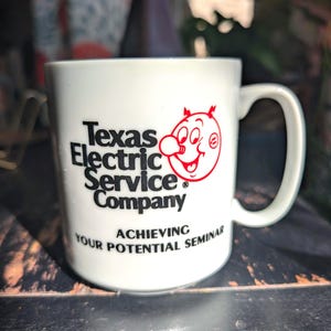 May include: White ceramic mug with the text "Texas Electric Service Company" in black, and a red cartoon character. The mug also has the text "Achieving Your Potential Seminar".