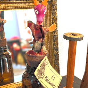 May include: A decorative jar with a wooden deer-like figure on top. The figure has a pink head, orange antlers, and a patterned fabric body. A tag reads "El Nahualito" with an illustration. The jar is brown and sits in front of a gold framed mirror.