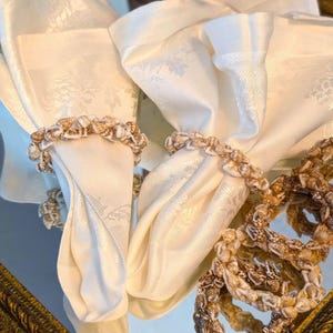 May include: Ivory-colored cloth napkins with a subtle floral pattern, paired with seashell napkin rings. The rings are made of small, natural seashells in shades of brown and cream, adding a coastal touch to the table setting.
