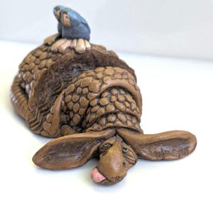 May include: A brown, textured armadillo figurine with a segmented shell and long ears, lying on its stomach with a small pink tongue visible. Two small, blue-grey bird figures are perched on the back of the armadillo's shell.
