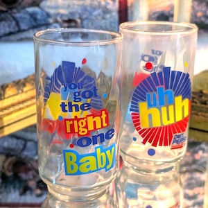 Puede incluir: Dos vasos de vidrio transparentes vintage con gráficos coloridos. Un vaso tiene el texto "you got the right one Baby" en azul, amarillo y rojo. El otro vaso tiene "uh huh" en amarillo y rojo. Ambos vasos se reflejan.