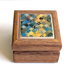 May include: A square wooden box with a visible grain texture. The lid features an inset square tile decorated with a colorful patchwork pattern of blue, yellow, and green floral and geometric shapes arranged in a diamond grid layout.
