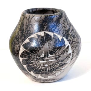 May include: A rounded ceramic vase featuring a smooth, light grey surface with intricate black horsehair-style markings. A circular medallion is etched into the centre, displaying a stylised sun face with radiating blades.