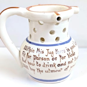 May include: A white ceramic puzzle jug with a handle and a perforated neck. The body features blue horizontal bands and brown hand-painted text that reads: Within this Jug there is good Liquor, Fit for Parson or for Vicar, But how to drink and not to spill, Will try the utmost of your skill.