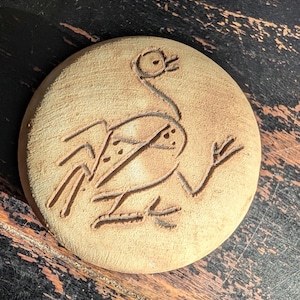 May include: A round, light brown ceramic coaster with a carved design of a bird. The bird has a long neck, a beak, and a stylized body with dotted details. The coaster is circular and has a slightly rough texture.