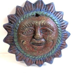 May include: A circular wall plaque shaped like a sun with a human face. The sun features a central face with a smiling expression, surrounded by a rope-like border and pointed rays. The finish is a distressed mix of rust red, blue, and green.