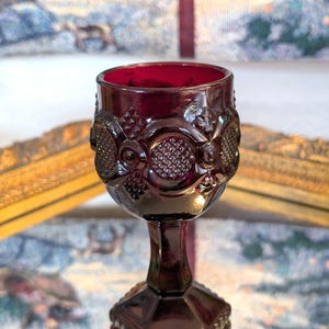Könnte beinhalten: Ein rubinroter Glasbecher im Vintage-Stil. Der Becher hat verzierte geprägte Details um die Schale und einen facettierten Sockel. Das Innere des Glases ist ein leuchtendes Rot, und der Stiel ist ein tiefes, sattes Rot.