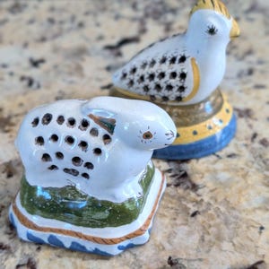 May include: Two ceramic animal figurines: a white rabbit and a bird. The rabbit has a series of holes on its back and sits on a green and blue base. The bird has a yellow beak and head, and sits on a blue and gold base.