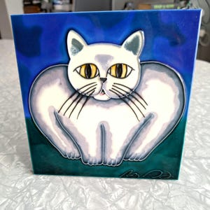 May include: Square ceramic tile featuring a stylized white cat with yellow eyes and black whiskers. The cat is set against a blue background with a green border. The artwork has a whimsical, cartoon-like style.
