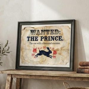 Puede incluir: Obra de arte enmarcada con el texto "WANTED: THE PRINCE." presenta una ilustración de un conejo negro. La obra se exhibe sobre un banco de madera, junto a una cesta tejida y un jarrón decorativo con vegetación.