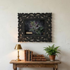 May include: A framed botanical print featuring a dark floral arrangement with purple flowers and black berries. The ornate black frame hangs above a wooden table with books, a brass lamp, and a potted fern. The print has a small text label.