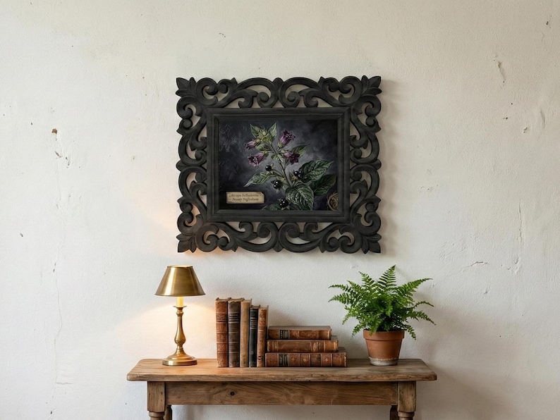 May include: A framed botanical print featuring a dark floral arrangement with purple flowers and black berries. The ornate black frame hangs above a wooden table with books, a brass lamp, and a potted fern. The print has a small text label.