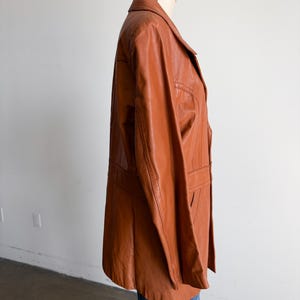 May include: A vintage, rust-colored leather jacket displayed on a mannequin. The jacket features a classic lapel collar, two front pockets, and a tailored fit. The jacket is paired with blue jeans.