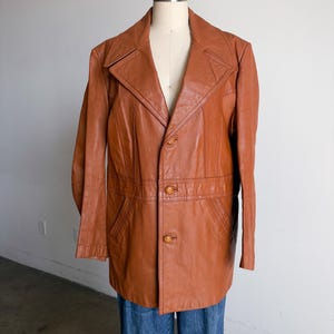 May include: A vintage brown leather jacket with a notched collar, two front pockets, and button closures. The jacket is displayed on a mannequin and paired with blue jeans. The jacket's length is mid-thigh.