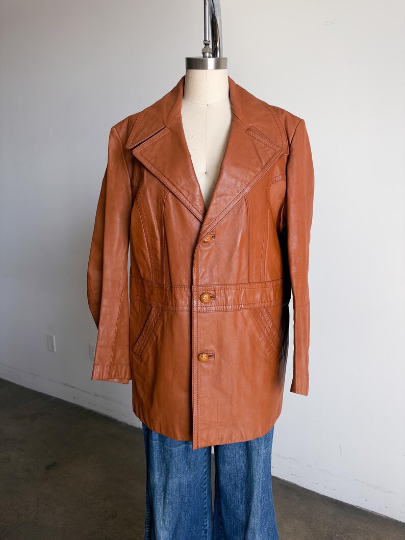 May include: A vintage brown leather jacket with a notched collar, two front pockets, and button closures. The jacket is displayed on a mannequin and paired with blue jeans. The jacket's length is mid-thigh.