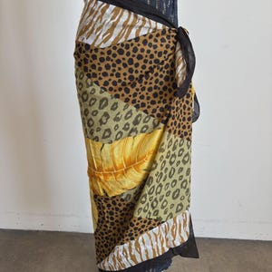 May include: A wrap skirt featuring a patchwork design of animal prints including leopard spots and zebra stripes, combined with a yellow tropical leaf pattern. The garment has a solid black waistband and a side tie closure with black fabric edges.