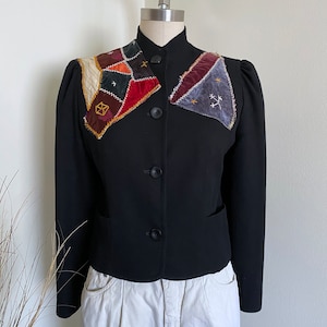 May include: A black cropped blazer with a unique patchwork collar featuring various colors and textures, including red, blue, green, and brown. The collar is adorned with gold embroidery.
