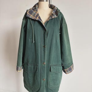 May include: A dark green hooded jacket featuring a button-front closure, two large front flap pockets, and long sleeves with cuffs. The interior lining and the underside of the hood and cuffs display a blue, yellow, and white plaid pattern.