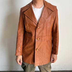 May include: A vintage brown leather jacket with a notched collar, three decorative buttons, and a belt detail at the waist. The jacket has a tailored fit and features pockets. The jacket is a medium brown color.