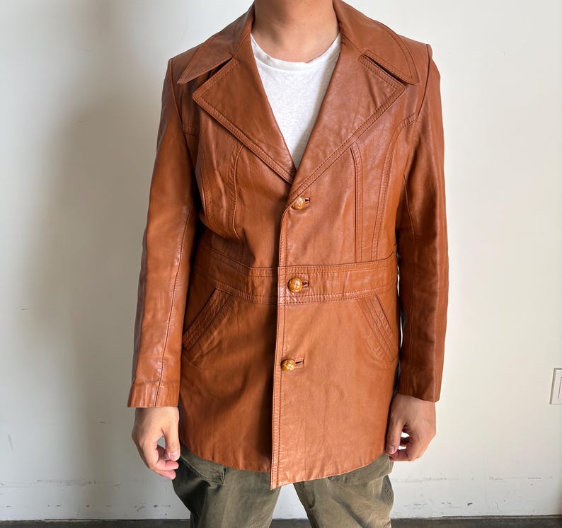 May include: A vintage brown leather jacket with a notched collar, three decorative buttons, and a belt detail at the waist. The jacket has a tailored fit and features pockets. The jacket is a medium brown color.