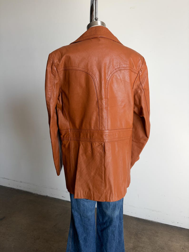May include: A vintage brown leather jacket with a notched collar and tailored fit. The jacket features detailed stitching and a classic design. The jacket is displayed on a mannequin with blue jeans.
