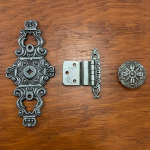 May include: Three silver-toned metal hardware pieces: a decorative hinge, a door knob, and a decorative plate with a crown on top.