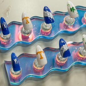 May include: A collection of artificial nails with diverse designs. Some nails are blue with silver detailing, others are white with a gold 'T', and some feature a green four-leaf clover. The nails are presented on a clear, iridescent display.