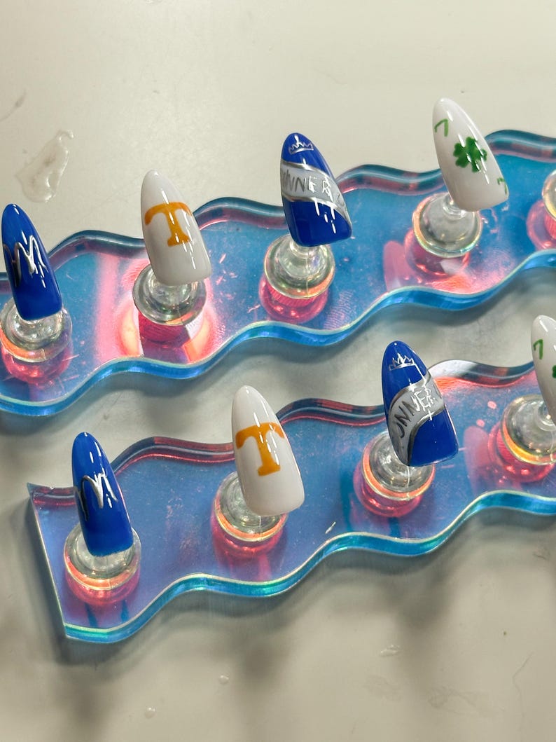 May include: A collection of artificial nails with diverse designs. Some nails are blue with silver detailing, others are white with a gold 'T', and some feature a green four-leaf clover. The nails are presented on a clear, iridescent display.