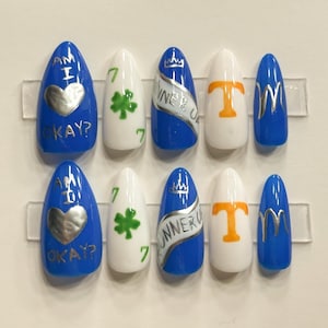 May include: A set of ten artificial nails in various designs. The nails are blue, white, and silver, with text and graphics including a heart, shamrock, and the words "WINNER UP".