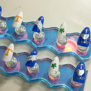 May include: A collection of artificial nails with diverse designs. Some nails are white with orange lettering, others are blue with silver text, and some are white with a green shamrock. The nails are presented on a clear, iridescent stand.