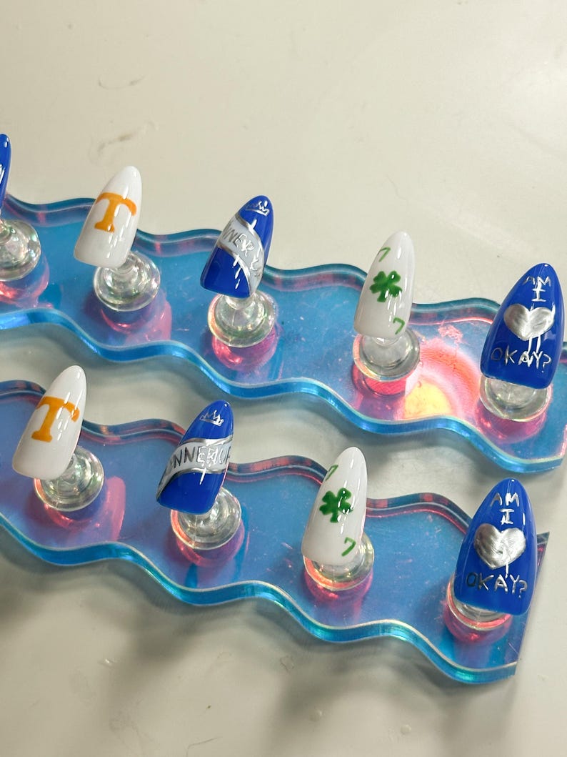 May include: A collection of artificial nails with diverse designs. Some nails are white with orange lettering, others are blue with silver text, and some are white with a green shamrock. The nails are presented on a clear, iridescent stand.