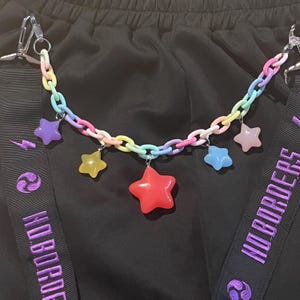 May include: A colorful chain with star charms in various colors, including red, yellow, purple, and blue. The chain is attached to black fabric with purple text that reads "NO BORDERS".