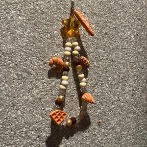 Könnte beinhalten: Ein Perlenanhänger mit Miniatur-Essensdekorationen. Der Anhänger enthält ein Baguette, Croissants, eine Waffel und andere Perlenakzente. Die Perlen sind mit einem Metallverschluss und einem sternförmigen Anhänger aufgefädelt.