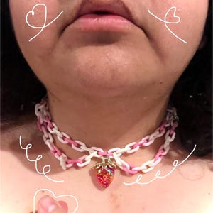 May include: A layered choker necklace with white and pink chain links. A strawberry pendant with red and gold accents hangs from the necklace. The necklace is worn around the neck.