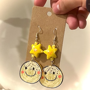 May include: Dangle earrings with a yellow star and smiling face design. The earrings have gold-coloured hooks and are attached to a brown card. The smiling face has a black outline and pink cheeks.
