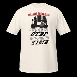 Op de afbeelding: Crèmekleurig T-shirt met de tekst "Breaking Boundaries" in het rood boven een zwart-wit afbeelding. De woorden "One Step at a Time" staan eronder in een verweerde letter.