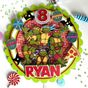 May include: A colorful cake topper featuring the Teenage Mutant Ninja Turtles. The topper has a red brick background with the number 8 and the name "RYAN". Decorative elements include ninja stars, pizza, and turtle shells.