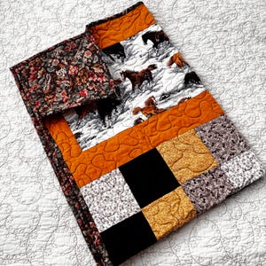 May include: A patchwork quilt featuring a horse-themed design. The quilt incorporates various fabrics, including a floral print, a horse scene on a white background, and solid orange and black squares. The quilt is a cozy home decor item.