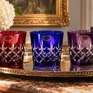 May include: Three crystal tumblers in red, blue, and purple, each featuring a diamond-cut pattern and a gold oval sticker near the rim. The glasses are arranged in a row, showcasing their vibrant colors and intricate, reflective glass facets.