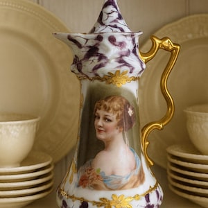 May include: A decorative porcelain pitcher with a portrait of a woman. The pitcher features a gold handle and floral accents. The body is white with purple and gold detailing. The pitcher is part of a tea set.