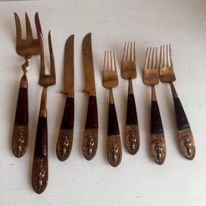 May include: A collection of antique cutlery, including forks and knives. The utensils have brass blades and tines, with dark red wooden handles. The handles are adorned with a decorative brass emblem.