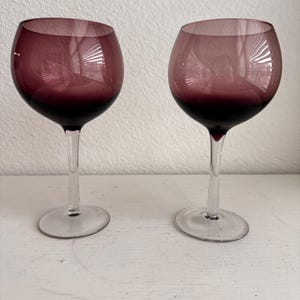 May include: Two wine glasses with a deep burgundy hue. The bowls transition from a lighter shade to a darker, almost black color at the base. The stems and bases are clear, creating a contrast with the colored bowls. The glasses are on a white surface.