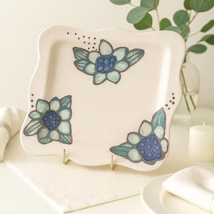 May include: A square, off-white ceramic plate with a wavy edge. The plate is decorated with hand-painted blue and teal floral designs. The plate is displayed on a gold stand, with a white napkin and candle in the background.