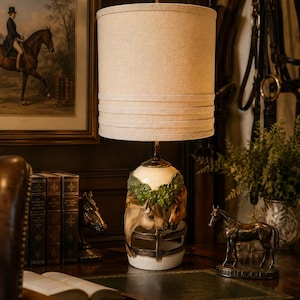 May include: A table lamp featuring a cylindrical beige fabric lampshade with horizontal stitched detailing. The ceramic base is decorated with three-dimensional horse heads emerging from a white fence structure with green foliage accents.