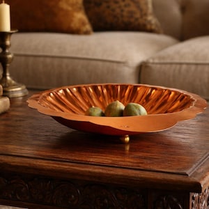 May include: A copper-colored decorative bowl with a fluted design, resting on a dark wooden coffee table. The bowl contains three green fruits. A candle holder and books are also on the table. The background includes a sofa.